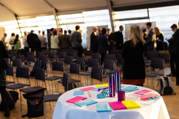 Personen stehen um Pinwände, auf denen sich Poster befinden und netzwerken. Im Vordergrund ist ein Stehtisch mit bunten Post-Its und Stifen