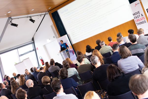 Vortragender auf dem Podium vor Publikum im Saal