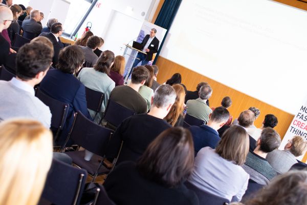 Vortragender auf dem Podium vor Publikum im Saal