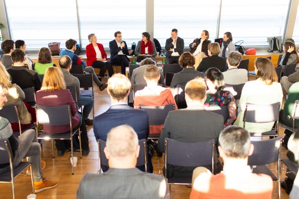 Sieben Personen auf dem Podium, davor sitzt das Publikum im Saal