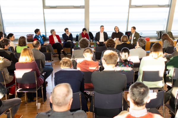 Sieben Personen sitzen in der Diskussionsrunde auf dem Podium, das Publikum sitzt halbkreisförmig darum herum