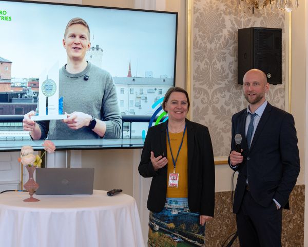 Mann mit Pokal auf dem Bildschirm, davor zwei Personen stehend